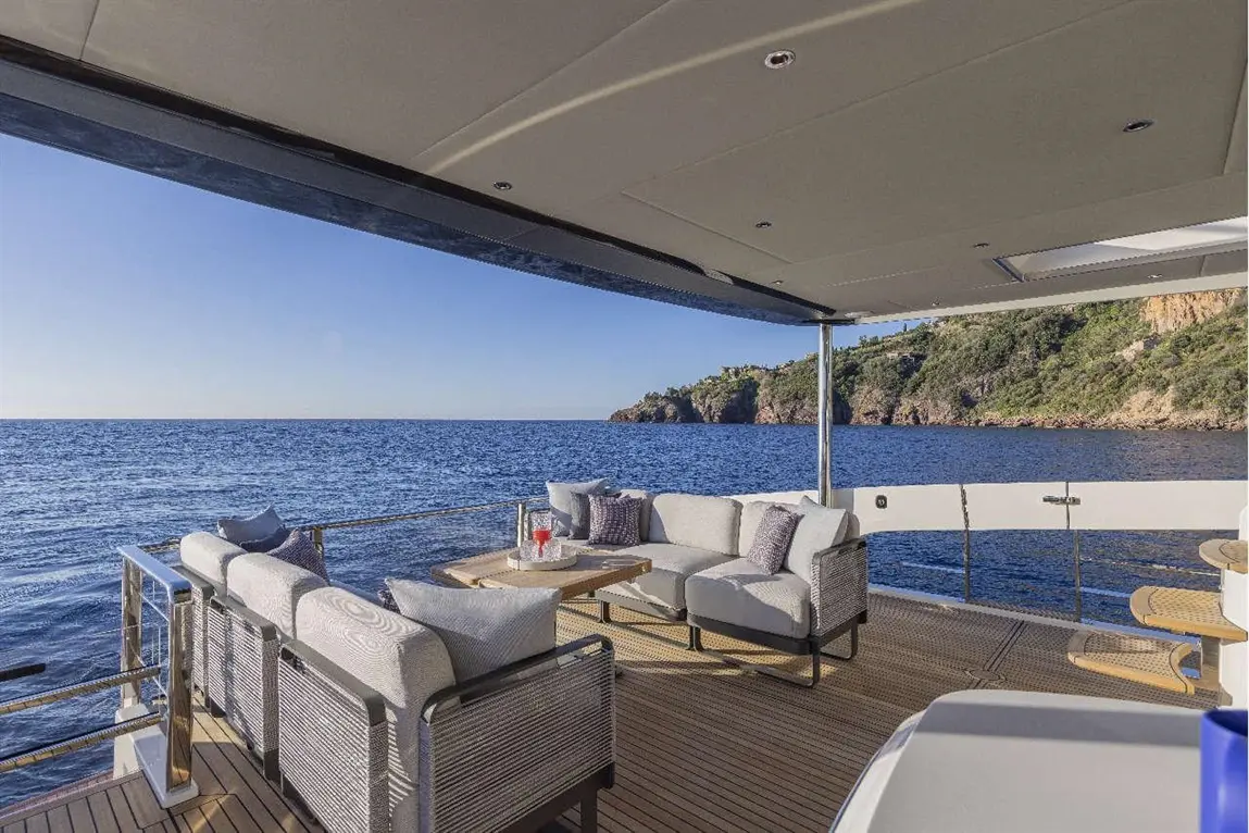 Luxury yacht deck with plush seating, a wooden table, and coastal views, overlooking calm blue waters and green hills.